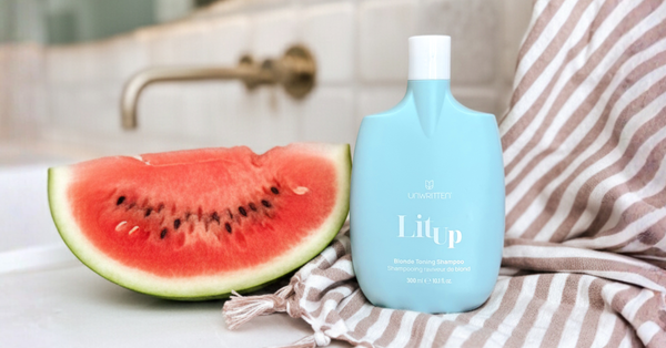 Watermelon slice beside Unwritten’s Lit Up Blonde Toning Shampoo on a bathroom counter.